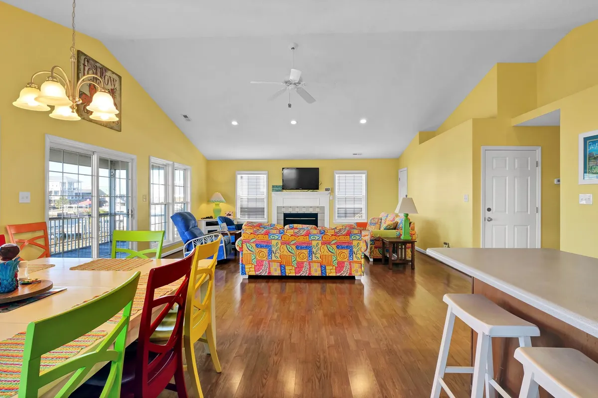 A bright living room scene featuring a colorful couch, a dining table with vividly painted chairs, and large windows bringing in natural light. The space is designed for socializing and relaxation, with a strong sense of warmth and connection and direct views out to the water.