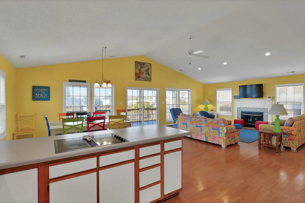 A spacious open-concept living area with colorful, inviting furniture arranged around a fireplace and large kitchen island. The room features large windows offering glimpses of the canal, playful artwork on the walls, and a cheerful, relaxed atmosphere perfect for gatherings, cooking, and evenings by the fire.