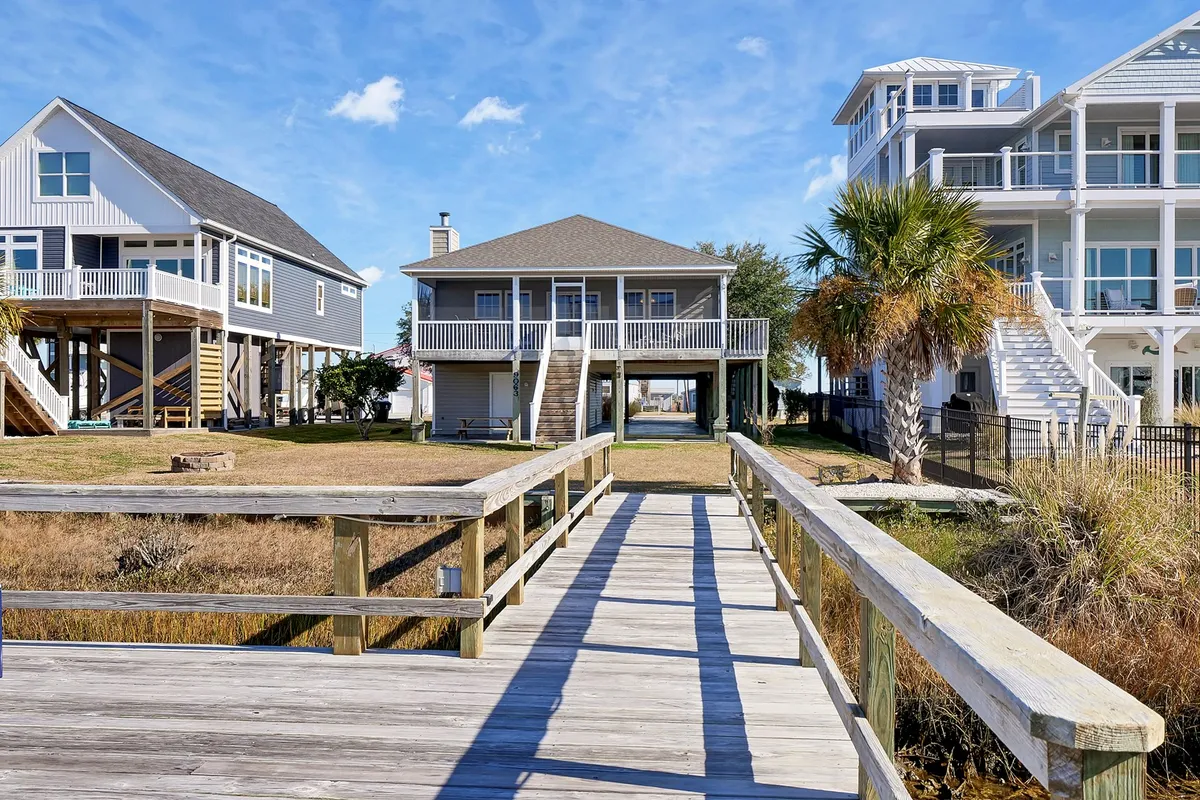 A wooden dock stretches out over the water, bordered by lush palm trees and flanked by two beach houses. The tranquil scene invites outdoor relaxation, fishing, and family gatherings, resonating with the vacation-home vibe and group outings.