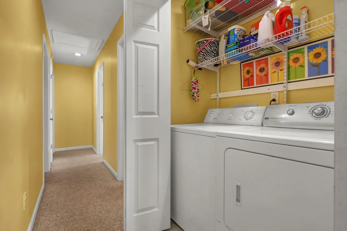A well-lit laundry area featuring a washing machine, shelves with organized cleaning supplies, and decorative wall art. The space is practical yet cheerful, supporting the idea of in-home convenience.