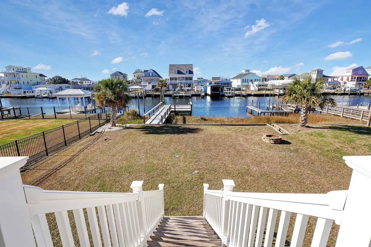 The image showcases the inviting backyard of a single-family house set along a tranquil canal in Surf City, North Carolina. Visible features include a lush lawn with a prominent fire pit surrounded by chairs, swaying palm trees, neighboring houseboats, and sparkling water perfect for boating or fishing. The scene exudes a cheerful, relaxed atmosphere ideal for outdoor gatherings.