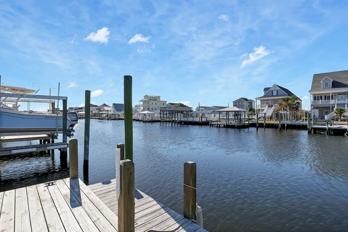 This tranquil waterside scene features a wooden dock jutting over calm canal waters, with boats tied alongside and neighboring houses in the background. The setting sun casts a warm glow, enhancing the inviting and peaceful atmosphere. The scene is ready for a group picnic, fishing session, or a quiet morning by the water.