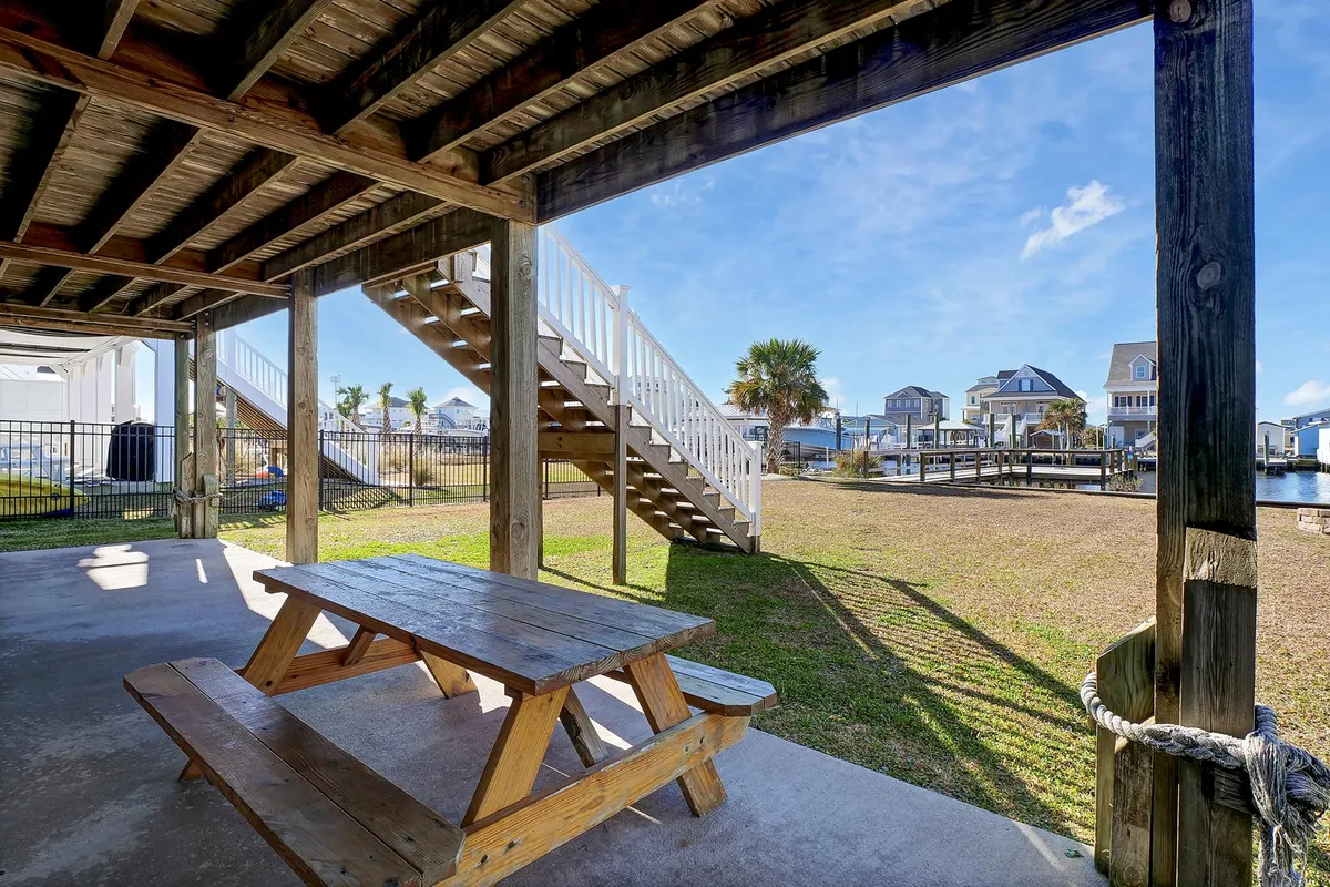 A cozy wooden picnic table sits near a staircase overlooking the tranquil canal, framed by palm trees and a fenced yard. The dock is visible, suggesting easy access to the water. The inviting setup is ideal for outdoor dining, morning coffee, or simply soaking up the peaceful ambiance by the canal.