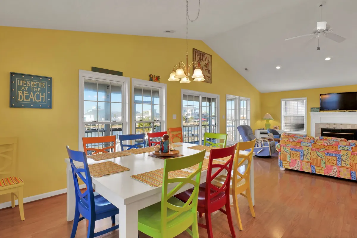 This image showcases a bright, lively interior room featuring colorful dining chairs, vibrant yellow walls, large windows letting in natural light, and a cozy couch with playful patterns. The space exudes a sense of energy and togetherness, ideal for gatherings, playing games, or relaxed downtime.