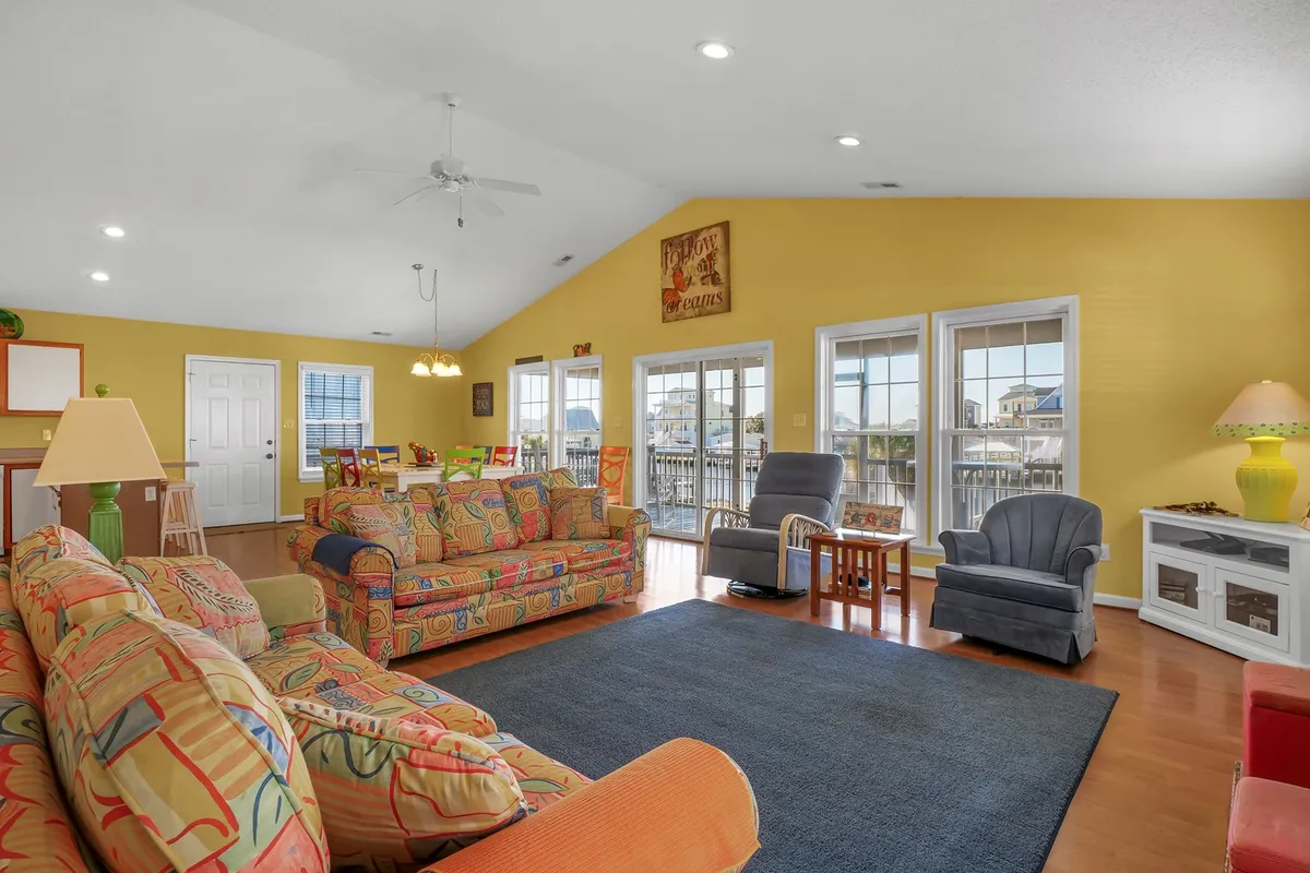 A spacious, inviting interior featuring colorful sofas, abundant natural light from large windows, a dining area, and warm wooden flooring. The room encourages group activities like playing games, socializing, or simply relaxing together.